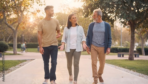 Tres personas de distintas edades caminando juntas por un parque urbano arbolado con luz cálida de atardecer