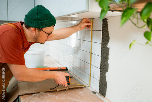 Foto Man wearing a green beanie measuring and affixing white ceramic tiles to a kitch