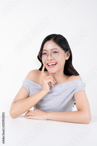 Cheerful young Asian woman smiling with finger touching cheek and wearing glasses on white background.