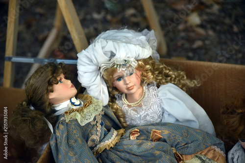 Two elegant retro dolls wearing beautiful vintage dresses displayed among old objects at a flea market, creating a nostalgic atmosphere around childhood and collectible toys.
