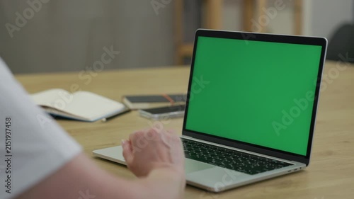 Woman Working On Laptop Computer with Green Mock Up Screen At Home Office Space. Female Freelancer Browsing Green Laptop Screen Writing Information in Notebook