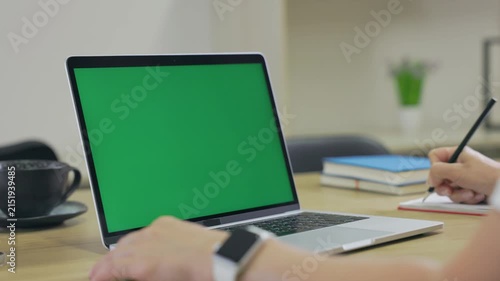 Woman Working On Laptop Computer with Green Mock Up Screen At Home Office Space. Female Freelancer Browsing Green Laptop Screen Writing Information in Notebook