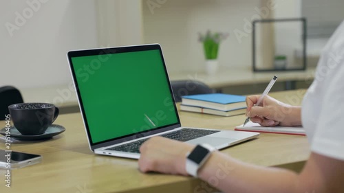 Woman Working On Laptop Computer with Green Mock Up Screen At Home Office Space. Female Freelancer Browsing Green Laptop Screen Writing Information in Notebook