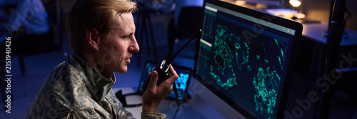 Caucasian young adult military officer speaking into walkie talkie while looking at computer screen with digital map. Image suitable for defense technology and security services marketing