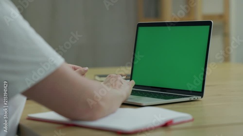 Woman Working On Laptop Computer with Green Mock Up Screen At Home Office Space. Female Freelancer Browsing Green Laptop Screen Writing Information in Notebook