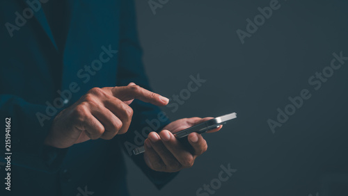 A person in a dark suit using a smartphone against a dark background, focusing on hand interaction with the device.