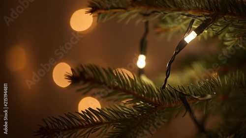 A close-up of Christmas tree branches with string lights twinkling on and off in a gentle sequence, soft pine needle texture visible, warm bokeh in the background, seamless loop.