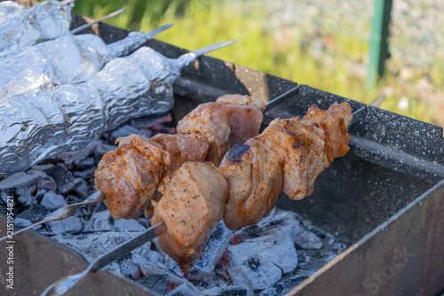 Delicious meat shashlik skewers and foil-wrapped food grilling on hot coals in a metal brazier outdoors. Summer picnic backyard barbecue cooking process with smoke and glowing charcoal embers.