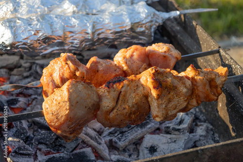 Delicious meat shashlik skewers and foil-wrapped food grilling on hot coals in a metal brazier outdoors. Summer picnic backyard barbecue cooking process with smoke and glowing charcoal embers.