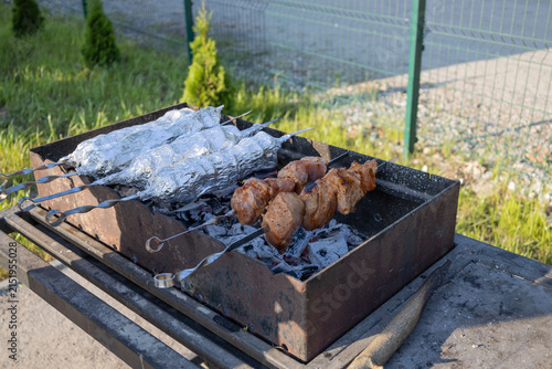 Delicious meat shashlik skewers and foil-wrapped food grilling on hot coals in a metal brazier outdoors. Summer picnic backyard barbecue cooking process with smoke and glowing charcoal embers.
