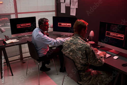 Middle aged Indian security guard and Caucasian soldier working on computers with critical error screens, managing cyber security breach. Useful for IT defense and crisis management ads