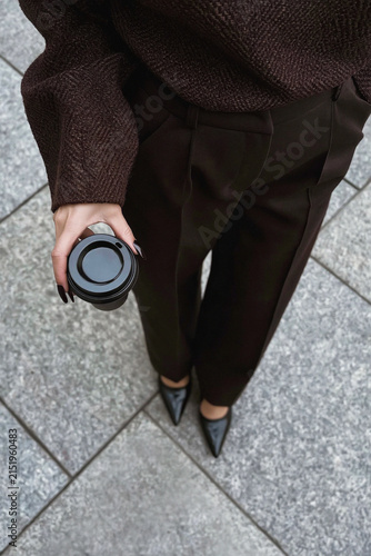 Top-down view of a stylish person in dark brown clothing holding takeaway coffee while standing on gray stone pavement.
