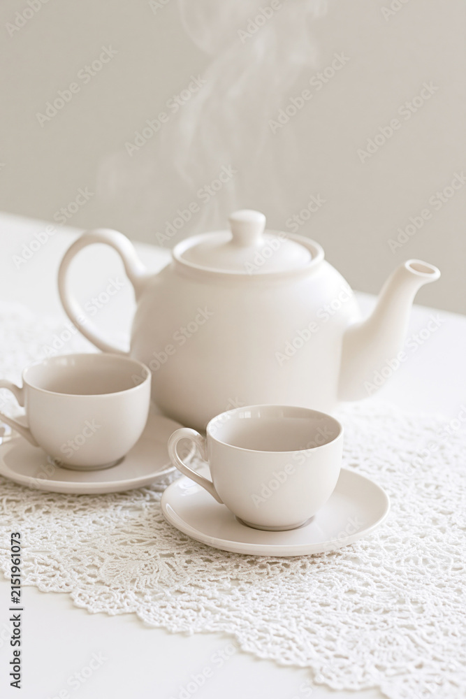 Fototapeta premium White ceramic teapot and two teacups rest on delicate lace as soft steam rises in a serene minimalist setting.