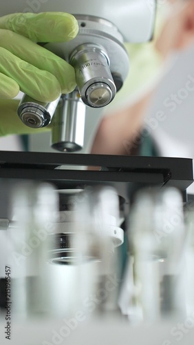 Close up of a scientist in green gloves and a mask, adjusting a microscope while conducting research in a lab with test tubes nearby. Medicine, healthcare and science concept