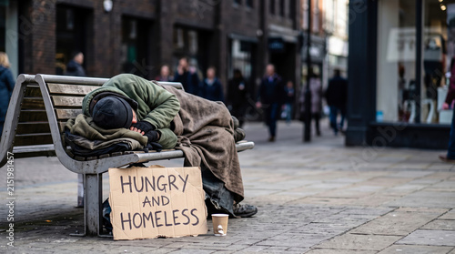 A person is sleeping on a bench in a city. They have a sign that says hungry and homeless next to them. People walk by in the background as they go about their day