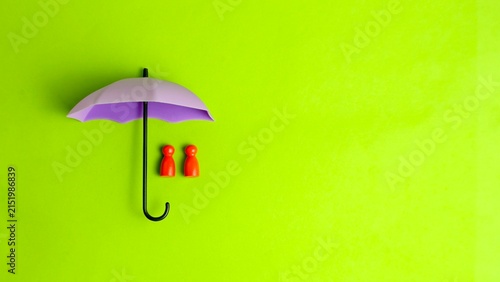 Purple miniature umbrella shielding two red wooden figures on a vibrant green background, representing family insurance protection.