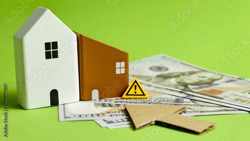 Wooden house models placed on US dollar banknotes with a warning sign and cardboard arrow pointing right on green background.