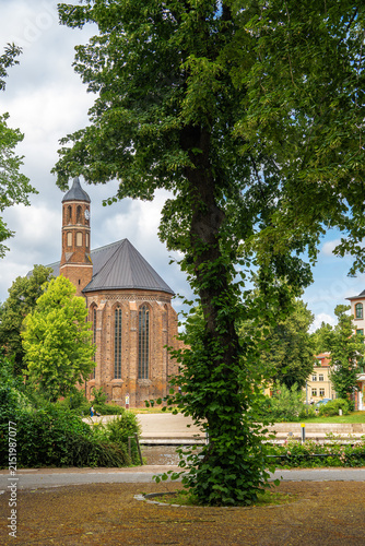 A large deciduous tree frames the view of a historic brick church with a clock tower, set in a green park under a changeable sky