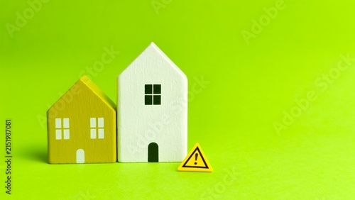 Yellow and white wooden house models displayed alongside a yellow triangle warning sign on a bright green background.