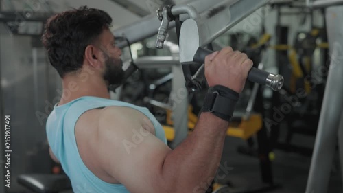 An Indian Asian fit young man using a lat pulldown machine, performing a resistance weight training exercise or workout targeting the back and shoulders in a modern gym. Fitness, bodybuilding concept