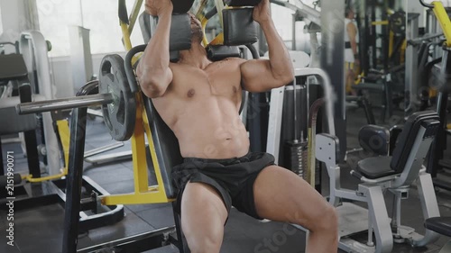 An Indian Asian fit muscular shirtless male performs a leg squat exercise using a machine as part of resistance or weight training in modern gym. Fitness, workout, healthy lifestyle, wellness concept