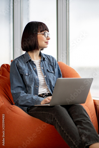 A woman with glasses and a bob haircut sits comfortably on an orange beanbag chair, typing on her laptop near a large window