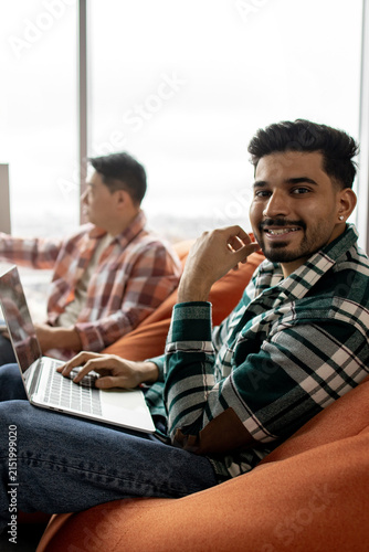 A smiling young man with a beard and dark hair is working on a laptop while sitting on an orange beanbag chair next to a window