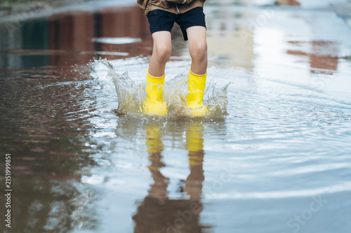 Boy in yellow rain boots splashes water in a summer puddle
