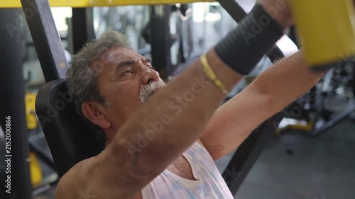 An Indian Asian fit healthy elderly or old man performs a resistance or weight training exercise using a machine in a modern gym. Fitness, workout, healthy lifestyle, wellness, bodybuilding concept
