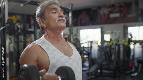 An Indian Asian attractive fit, muscular elderly man performs a single-arm dumbbell bicep curl exercise in modern gym. Fitness, resistance workout, weight training, healthy lifestyle, wellness concept