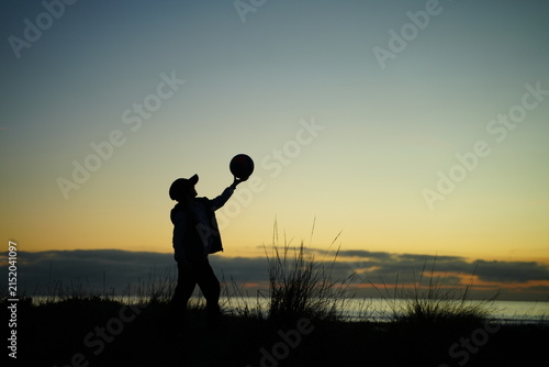Silhouette of a child balancing a ball on one finger against a dramatic sunset backdrop, highlighting playfulness and freedom. The unrecognizable figure adds a sense of mystery.