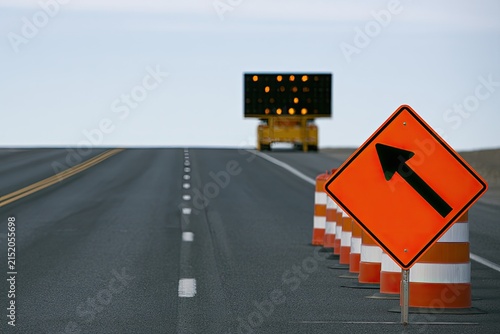 Orange directional sign and traffic cones lead down an empty road toward a lit arrow board