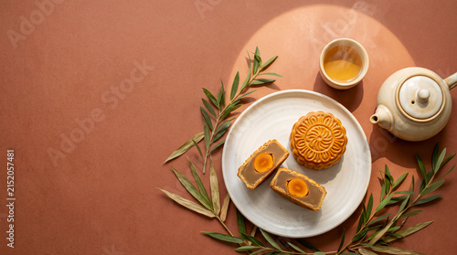 Traditional Mooncake Delight: A close-up view showcases a whole mooncake and a halved mooncake with golden yolk filling, artfully arranged on a white plate with a steaming cup of tea and a teapot.