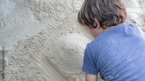 A young boy shapes and smooths a sand sculpture while playing outdoors in a tropical setting. The close-up captures creativity, imagination, and hands-on childhood exploration through natural play.
