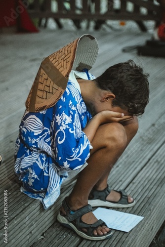Child wearing a traditional blue kimono and paper hat sits with head in hands, appearing thoughtful. The image reflects cultural attire and evokes a sense of introspection and calm.