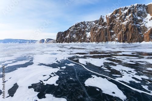 Winter landscape of frozen Baikal Lake with snow-capped mountains of Olkhon Island and white crusts of snow on blue ice surface on a cold, frosty February day. Winter ice travels