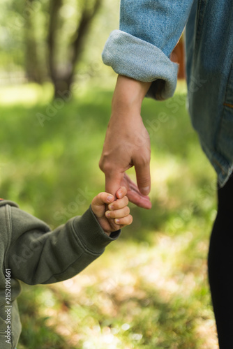 Small toddler holding mothers finger while walking outdoors in park.
