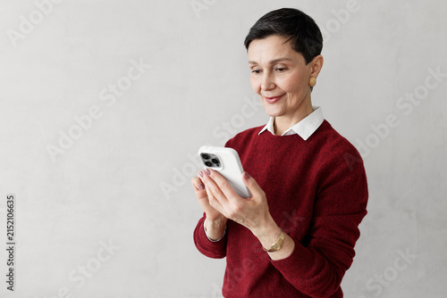 Middle-aged stylish woman entrepreneur in red jumper chatting with ai via smartphone application, using prompts to write content for social media promotion isolated on gray copy space background