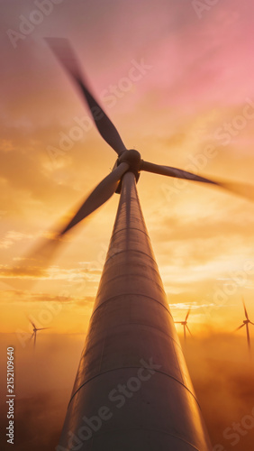 Low angle view of wind turbine tower rising into foggy orange sunset sky with motion blur.