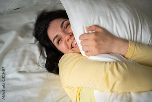 Smiling woman embracing pillow in cozy bed with soft bedding. Relaxation home comfort and emotional wellbeing concept