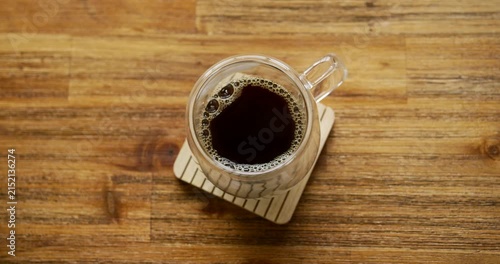 Placed clear glass mug of black coffee is settling on wooden coaster, bubbles shifting clockwise