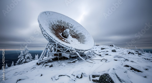 A large snow-covered satellite dish antenna perched atop a remote, winter-clad mountain peak under a dramatic sky