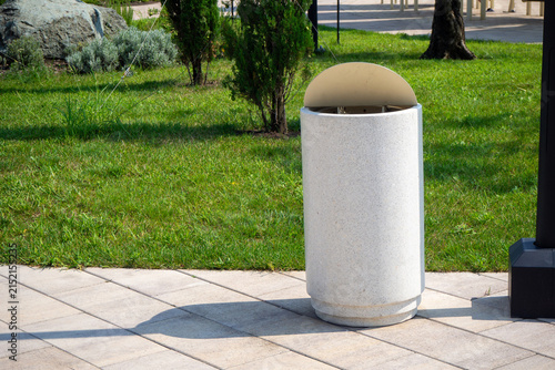 Konstfotografi White concrete cylindrical trash bin on paved sidewalk next to green lawn in park