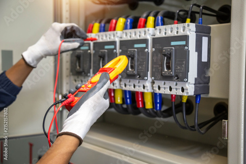 Obraz w ramie Industrial electrician holding digital clamp meter and using test probe to inspect commercial circuit breaker panel.