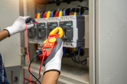 professional industrial electrician hands holding digital clamp meter and configuring test probes inside commercial circuit breaker panelboard.