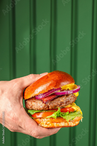 Hand holding a gourmet hamburger with lettuce, tomato, onion, and mustard against a green wooden background, showcasing a delicious meal presentation