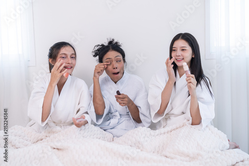 Obraz na płótnie Three young multiethnic women in soft white waffle bathrobes patting skincare cream onto their cheeks, as the woman with a long brown ponytail presses a fingertip of cream to her nose