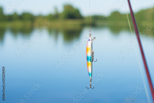 Colorful fishing lure hanging on line over calm lake with blurred shoreline. Ideal for sport fishing, lure selection, bait advertising, outdoor leisure and angling equipment promotions.