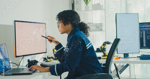 Young African American female robotics engineer coding AI software for robotic arm in laboratory. Embedded system, firmware testing, machine learning, industrial automation, future technology concept.