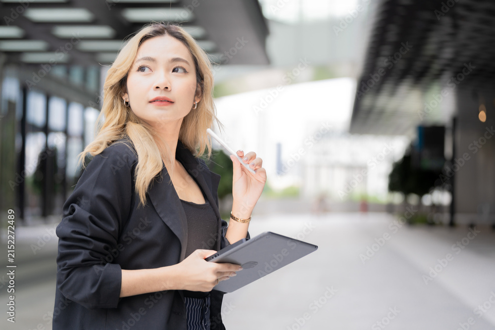 Fototapeta premium Asian businesswoman with long blonde-dyed wavy hair looks upward in thought with a stylus resting near her chin, tablet in hand along a covered walkway in Bangkok's business district.